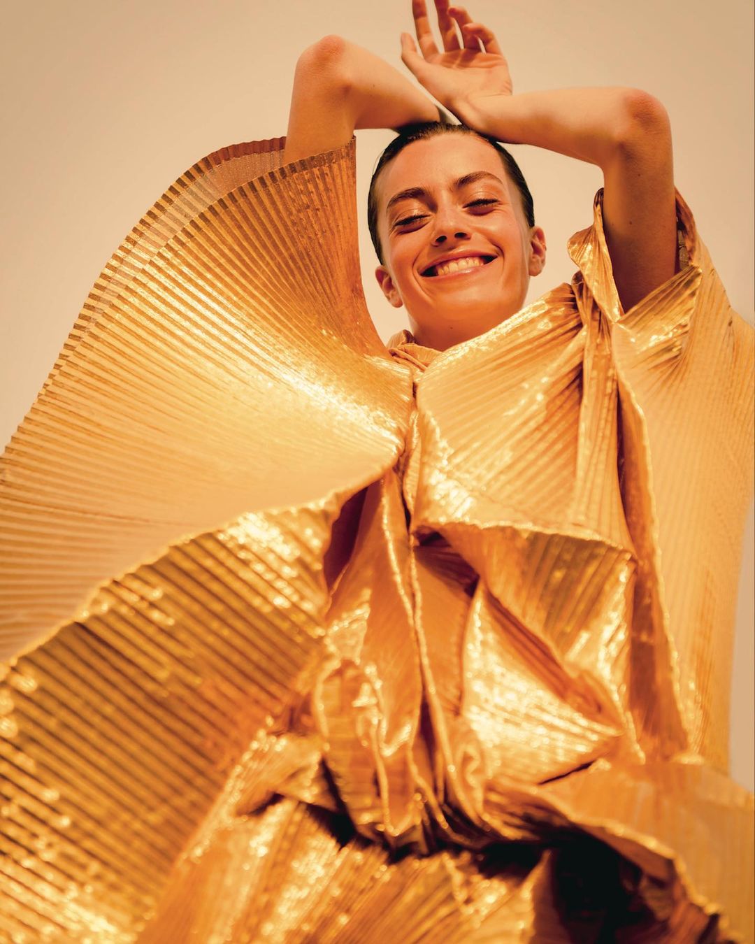 Smiling woman in a gold pleated top with her hands above her head in a studio setting with an off-white backdrop.