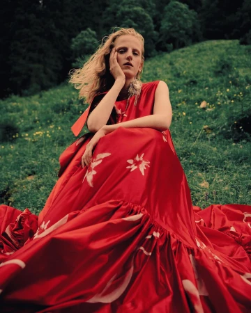 Model with long blonde hair seated outdoors on a grassy hill wearing a vibrant red dress with floral patterns.