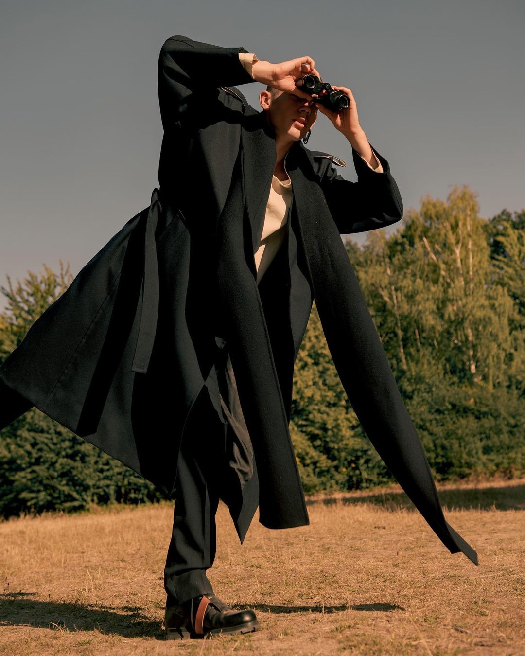 An adult male stands in a field with trees in the background, wearing a long, dark coat, looking through binoculars on a sunny day.
