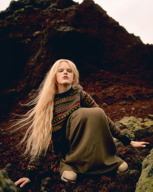An adult woman with long blonde hair poses seated on dark red volcanic rocks wearing a patterned knit sweater and a long brown skirt outdoors.