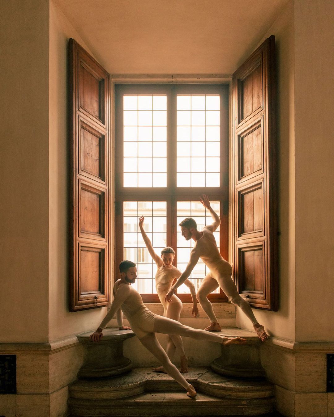 Three ballet dancers in light bodysuits pose in front of an open window with wooden shutters, showcasing a blend of art and architecture.