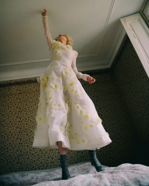 Woman in long, floral-patterned white dress and gray boots stands on a bed, reaching up toward the ceiling in an indoor room setting.