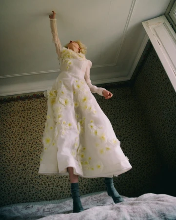 Woman in long, floral-patterned white dress and gray boots stands on a bed, reaching up toward the ceiling in an indoor room setting.