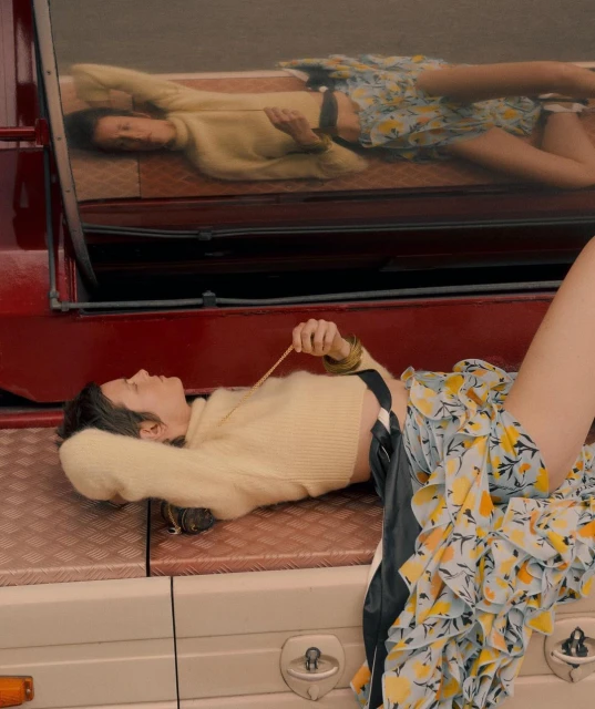 Adult female model poses on a red vehicle in a light-yellow crop top and floral skirt, reflected in the vehicle's window.
