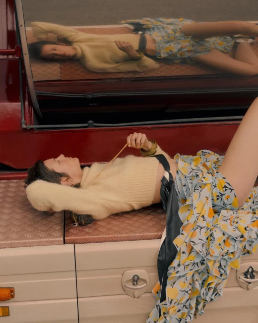 Woman in a yellow sweater and floral skirt rests on a red and white vehicle, reflected in the mirror, in a fashion editorial.