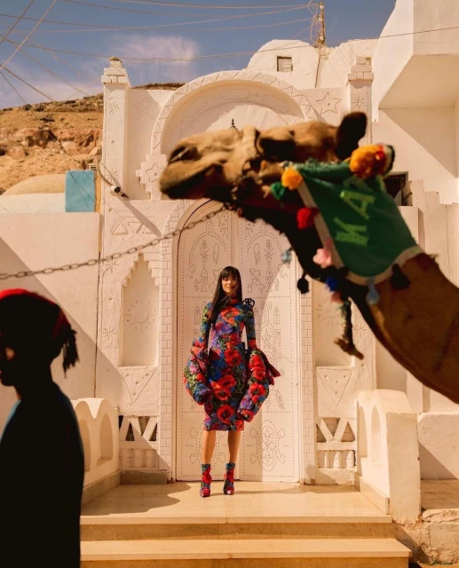 Woman in floral dress poses with camel and traditional architecture in bright daylight in an outdoor setting with warm lighting.