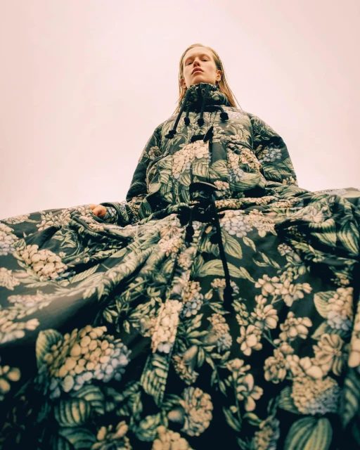 Eye-level shot of a single model wearing a floral print puffer jacket dress with a dark-tie detail against a pale pink sky background.