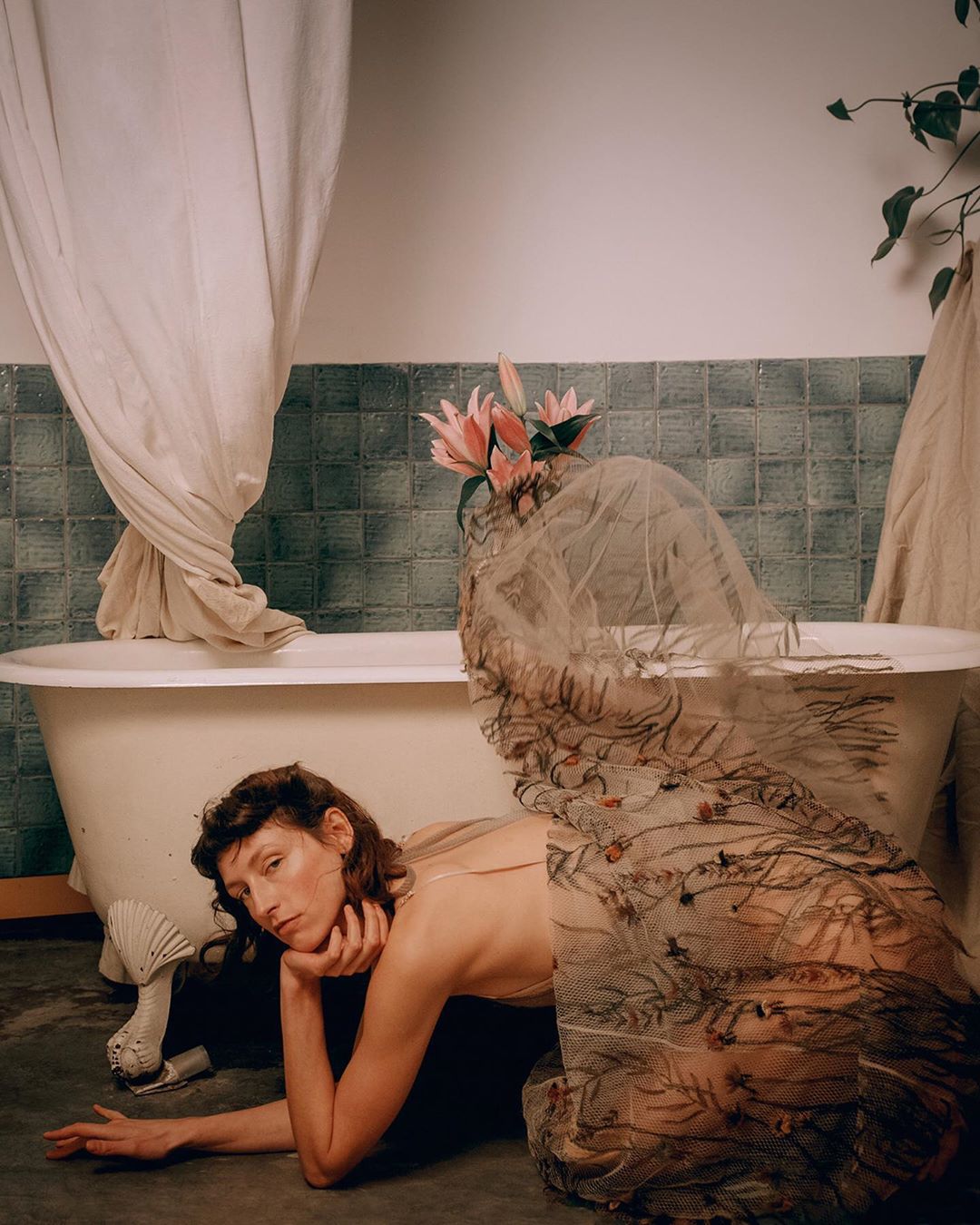 Woman in sheer floral dress lying on floor beside a bathtub in a tiled bathroom, photographed by Noemi Ottilia Szabo.