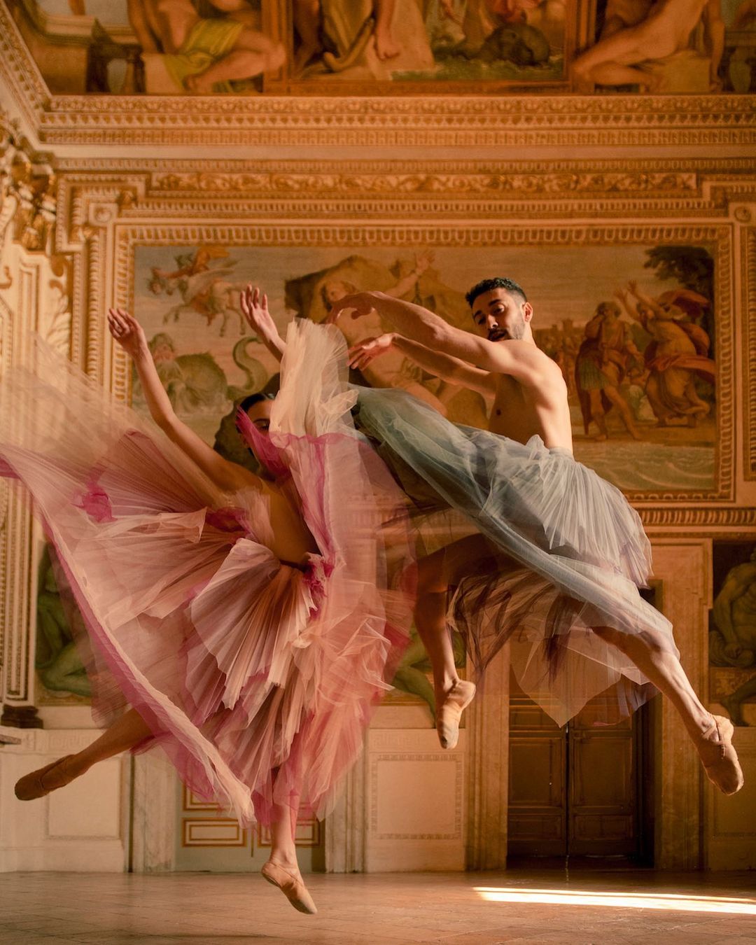 Dynamic ballet performance featuring a male and female dancer in mid-air wearing pink and grey tutus against a mural backdrop.