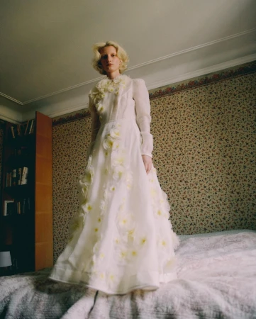 Model with blonde hair wearing a white long-sleeve floral applique dress standing in a bedroom