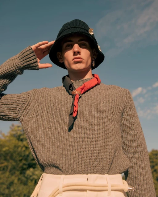 Eye-level portrait of an adult male in a knitted hat and sweater, saluting with a raised hand against a bright blue sky.