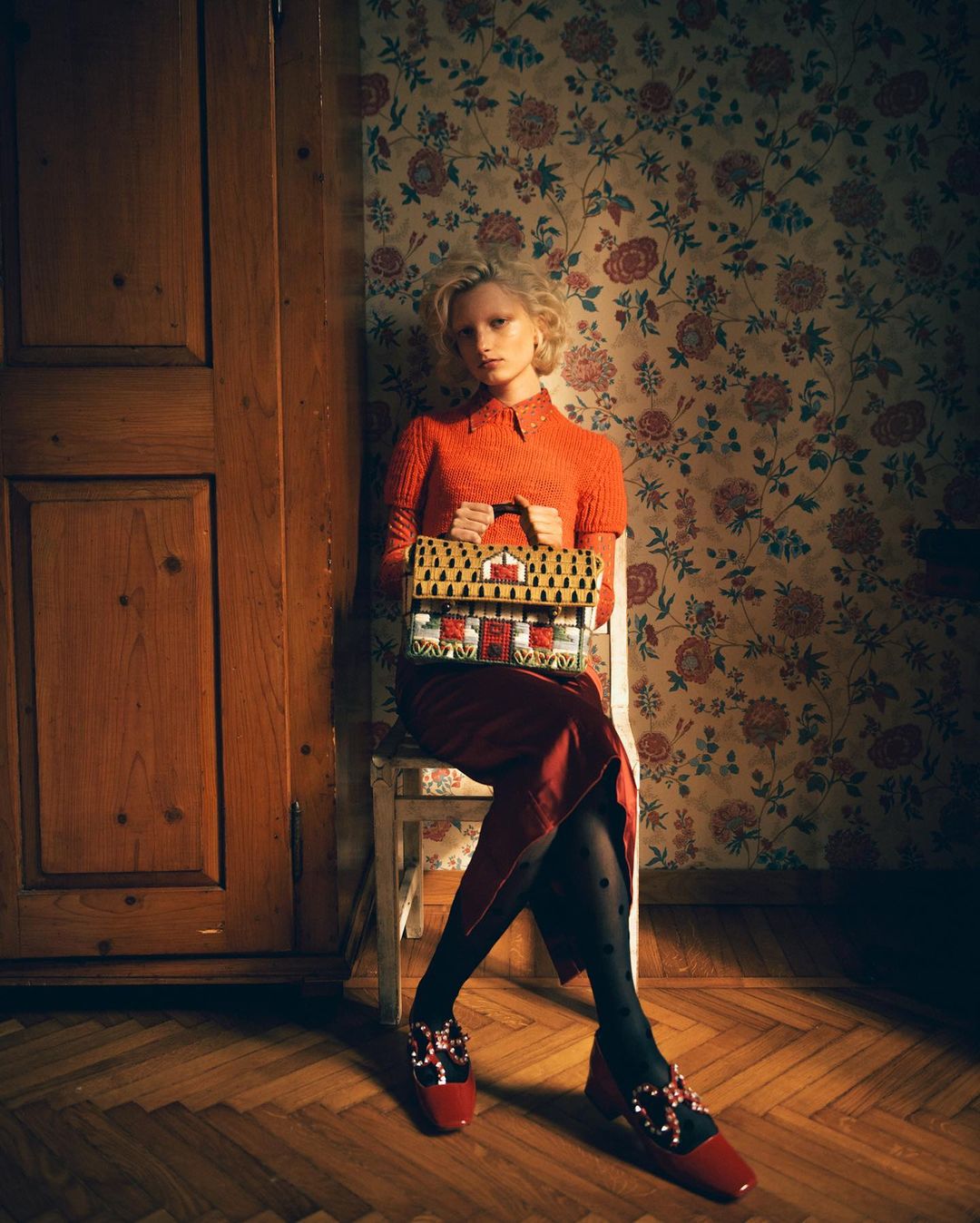 Indoor fashion shot of a blonde woman sitting on a chair, holding a handbag shaped like a house, red shoes, and a floral background.