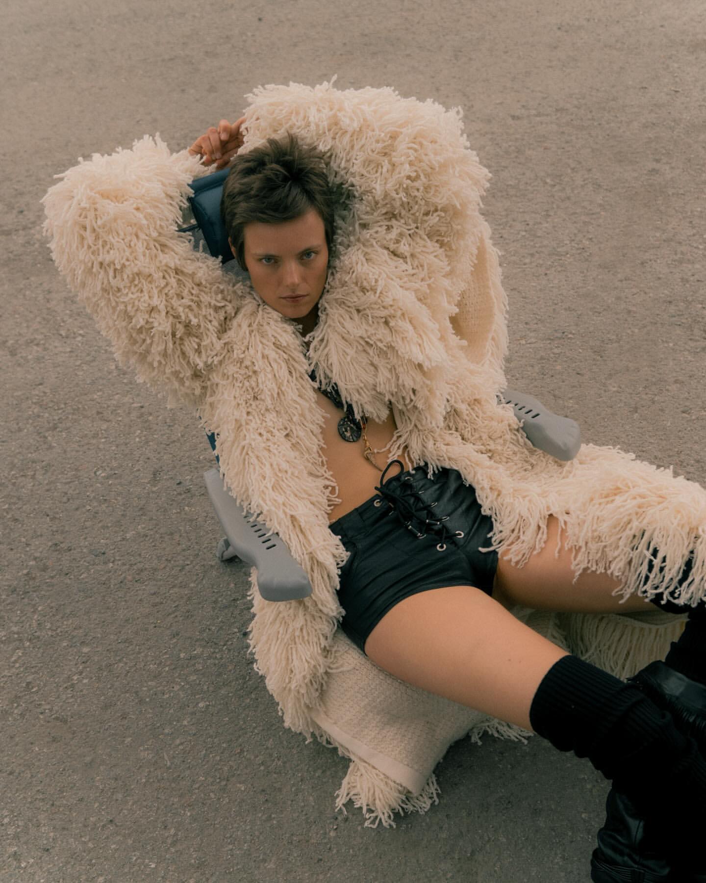 Person lies on a chair outside while wearing black shorts, boots, jewelry, and a shaggy cream coat in a fashion editorial.