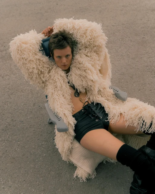 Person lies on a chair outside while wearing black shorts, boots, jewelry, and a shaggy cream coat in a fashion editorial.