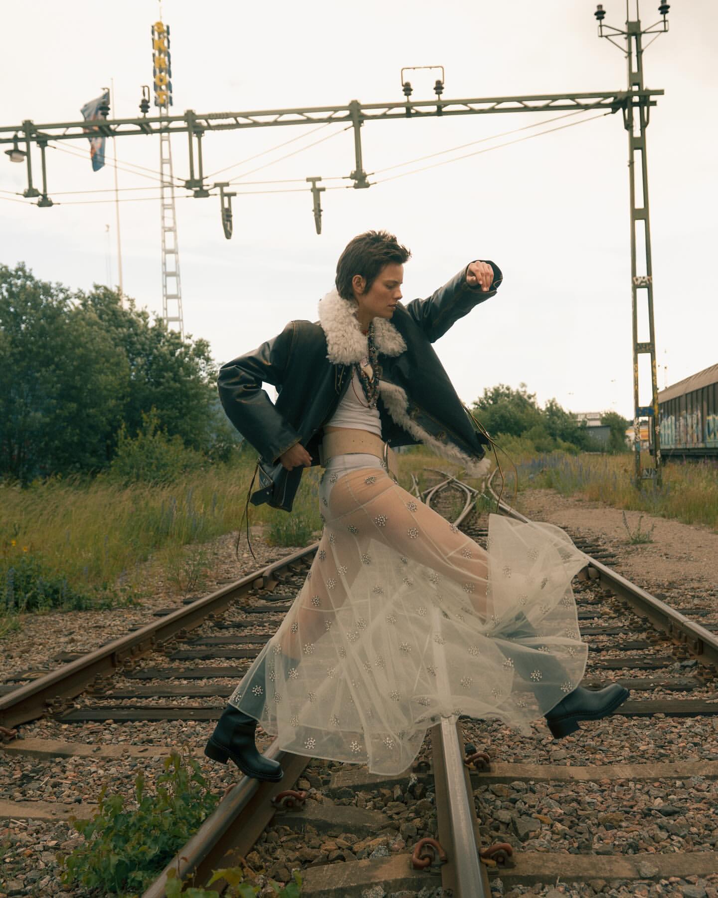 Woman in a sheer skirt and shearling jacket walking on railroad tracks with vegetation and an electrical tower in the background.