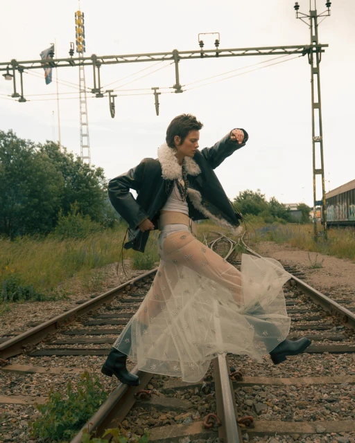 Woman in a sheer skirt and shearling jacket walking on railroad tracks with vegetation and an electrical tower in the background.
