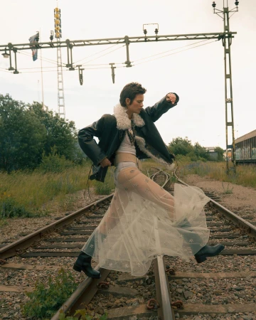 Woman in a sheer skirt and shearling jacket walking on railroad tracks with vegetation and an electrical tower in the background.