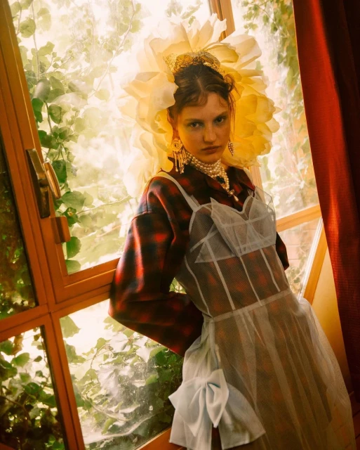 Portrait of a young woman in a plaid jacket and white dress with tulle, posing indoors near a window with a large floral headdress.