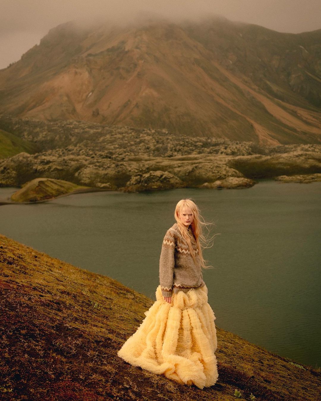 Young woman in grey sweater and yellow gown poses on a grassy hill with a still lake and misty mountains in the background.