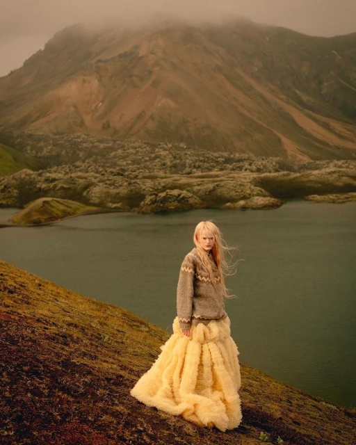 Young woman in grey sweater and yellow gown poses on a grassy hill with a still lake and misty mountains in the background.