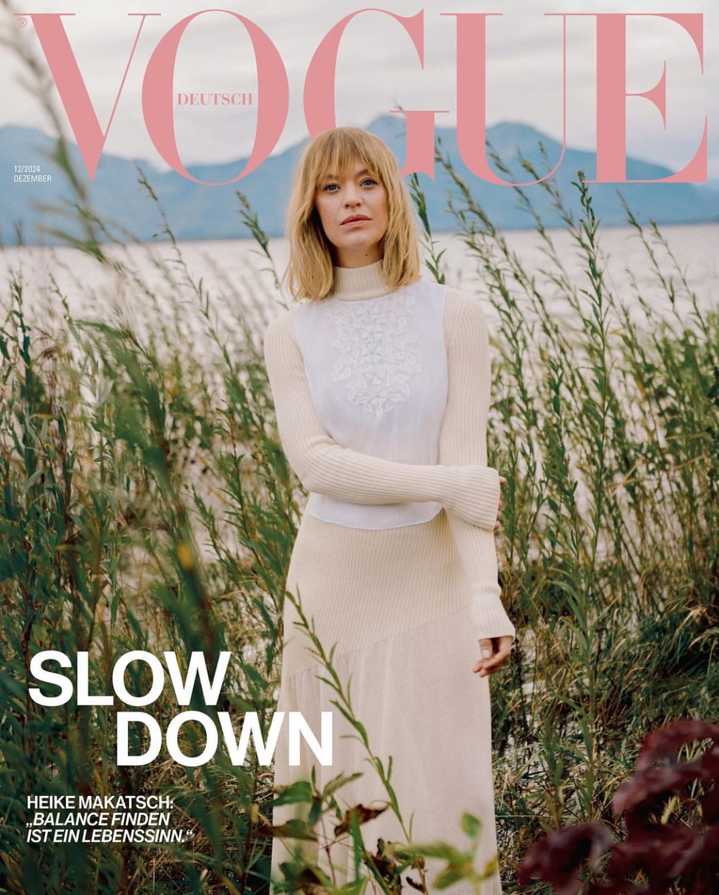 Heike Makatsch on the cover of Vogue Deutsch wearing a cream turtleneck sweater standing among tall grass by a lake.