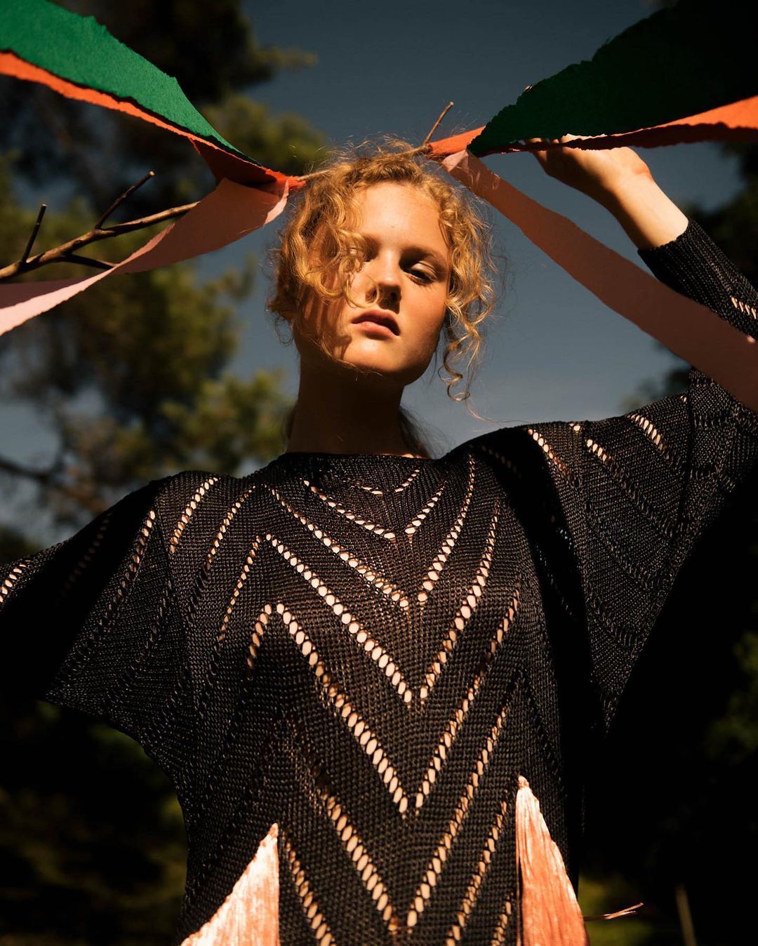 Adult woman with curly blonde hair poses outdoors in a black, fringed knit dress holding colorful ribbons, with a forest in the background.