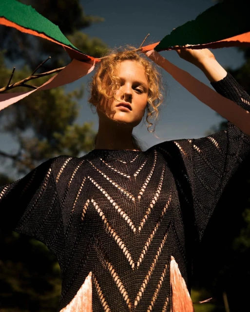 Adult woman with curly blonde hair poses outdoors in a black, fringed knit dress holding colorful ribbons, with a forest in the background.