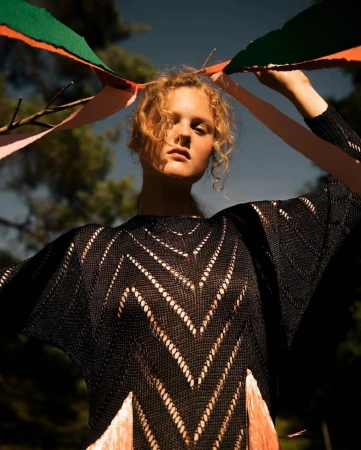 Adult woman with curly blonde hair poses outdoors in a black, fringed knit dress holding colorful ribbons, with a forest in the background.