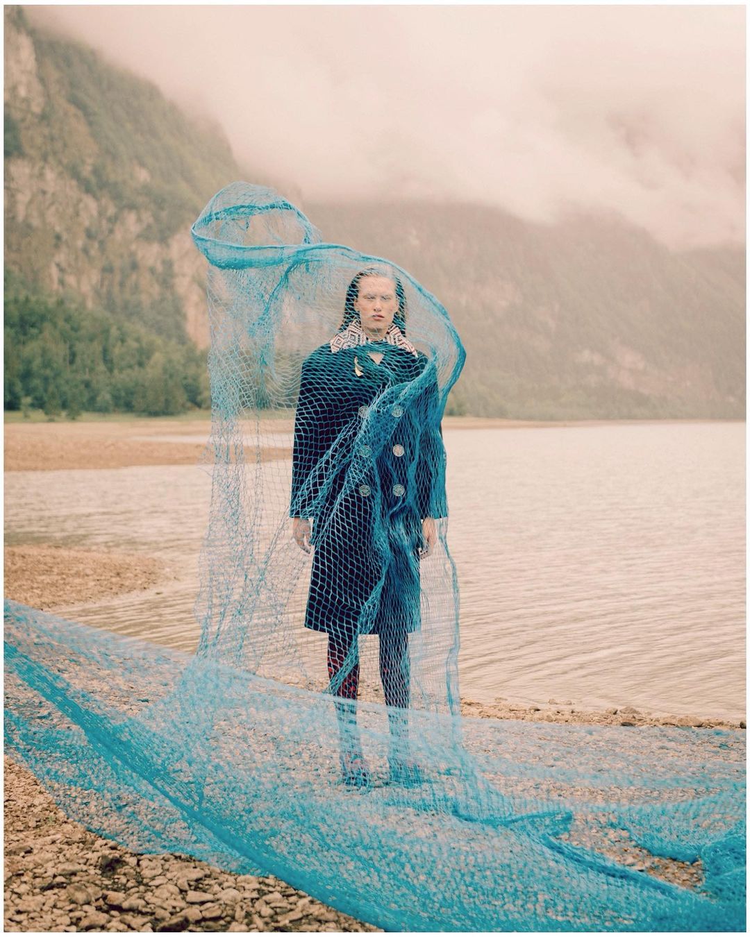 A woman stands on a shoreline with a blue fishing net, wearing a dark coat. Mountains and water are visible in the background.
