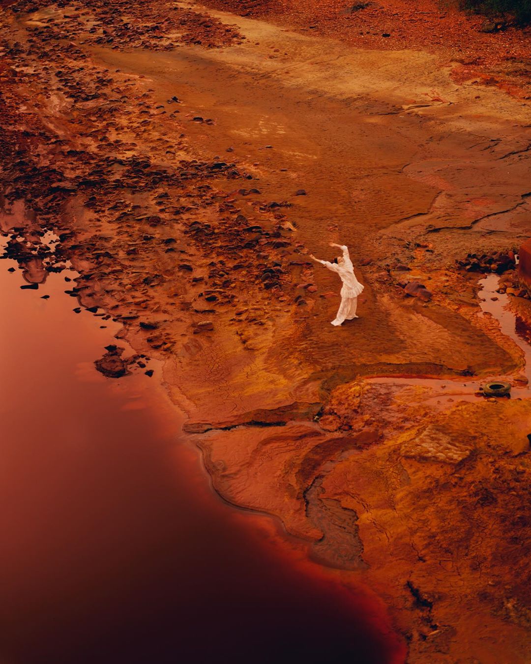 Woman in flowing white dress dances on red earth near red water, creating a stark and surreal landscape, viewed from above.