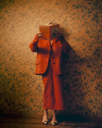 Person wearing an orange suit holding a book in front of their face against floral patterned wallpaper.