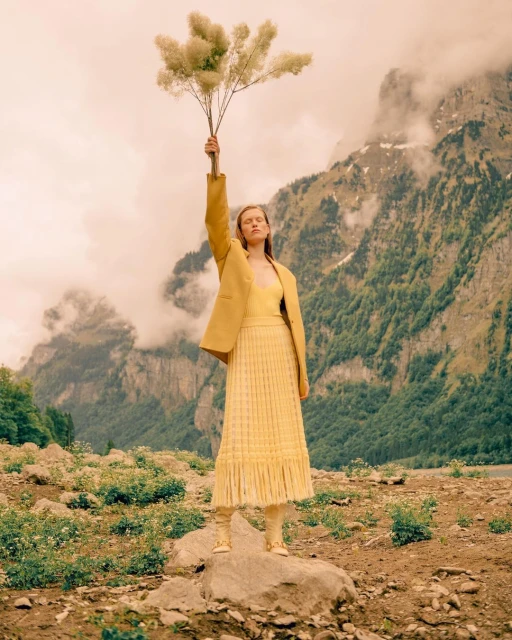 A young woman in a yellow suit stands on a rock holding up a large plant against a mountainous background with low-hanging clouds.
