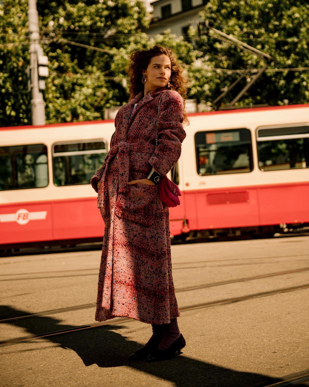 Woman in patterned red tweed coat and dark shoes standing on a city street with a red tram in the background.
