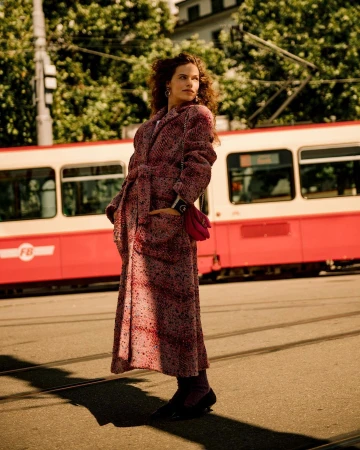 Woman in patterned red tweed coat and dark shoes standing on a city street with a red tram in the background.