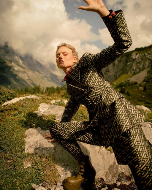 Fashionable woman in patterned suit poses on a rocky outcrop in mountainous landscape with cloudy sky, captured in low-angle shot.