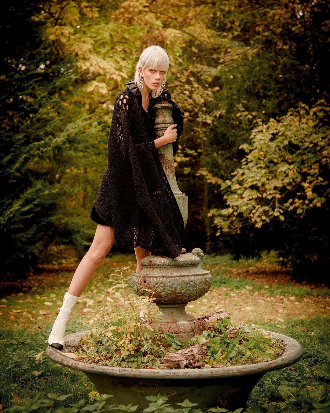 A woman with short blonde hair models a long black crocheted coverup and ivory boots in a park by a concrete fountain.