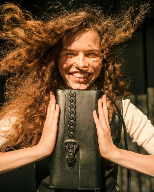Smiling model holding a black leather handbag with a chain detail. Fashion shot with bright lighting and curly red hair.