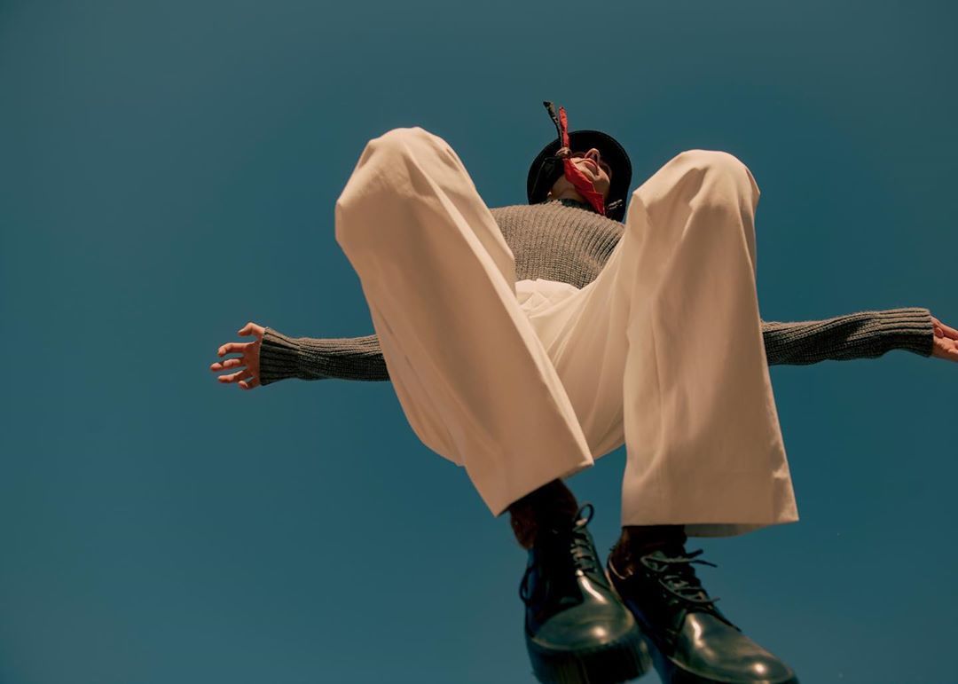 Eye-level view of a stylish adult wearing a hat, gray sweater, and white pants with shiny black shoes against a blue sky backdrop.