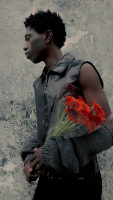 Black male fashion model in a vest poses with a bouquet of red flowers against a neutral textured background in a vertical portrait.