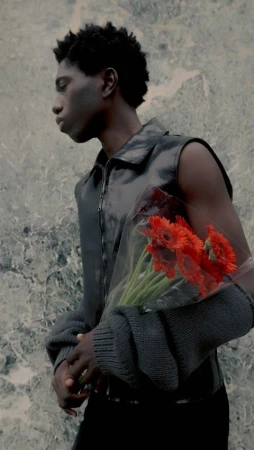 Black male fashion model in a vest poses with a bouquet of red flowers against a neutral textured background in a vertical portrait.