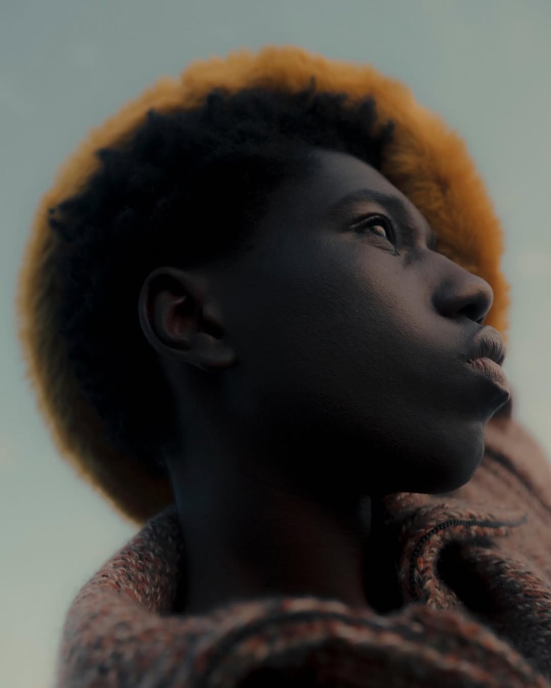 Close-up portrait of a young Black person with dark hair and blonde highlights, looking up with a soft gaze against a blurred background.
