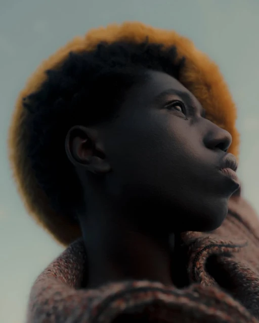 Close-up portrait of a young Black person with dark hair and blonde highlights, looking up with a soft gaze against a blurred background.