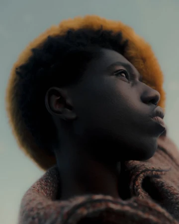 Close-up portrait of a young Black person with dark hair and blonde highlights, looking up with a soft gaze against a blurred background.