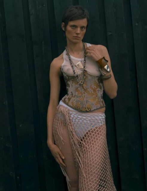 Woman with short hair poses in a halter top, corset, and net skirt against a dark wooden backdrop. She wears jewelry and wrist cuffs.