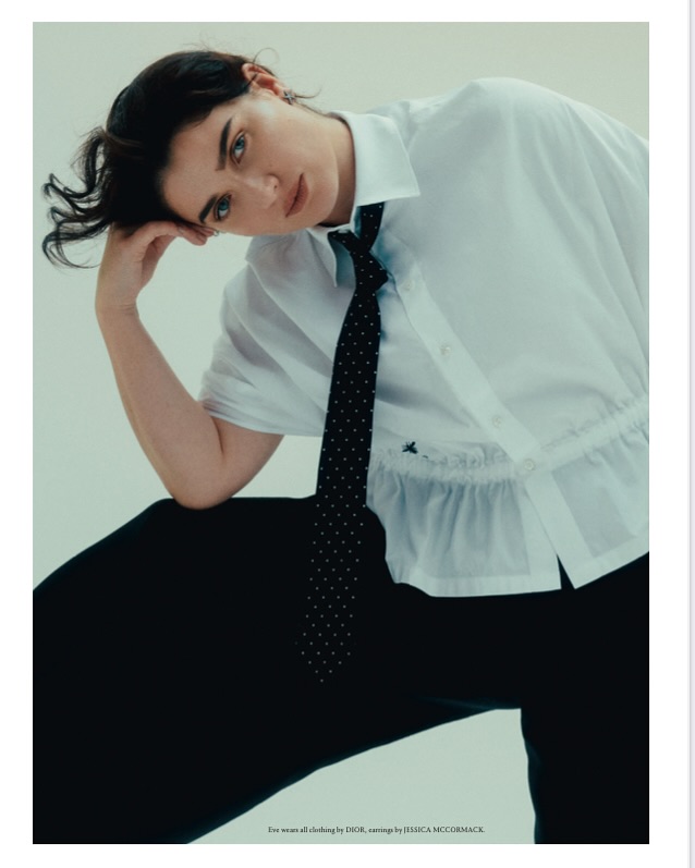 Portrait of a stylish young woman with dark hair wearing a white collared shirt and black tie, posing in front of a plain background.