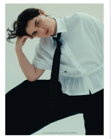 Portrait of a stylish young woman with dark hair wearing a white collared shirt and black tie, posing in front of a plain background.