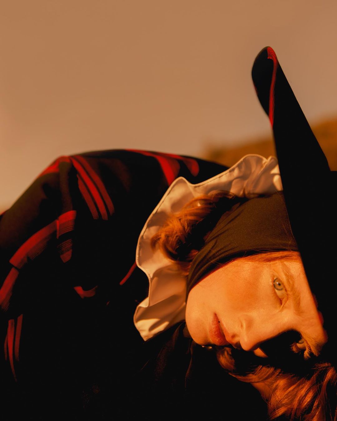 Portrait of adult woman with red hair wearing black bonnet with white frill and striped clothing lying on the ground in soft light.