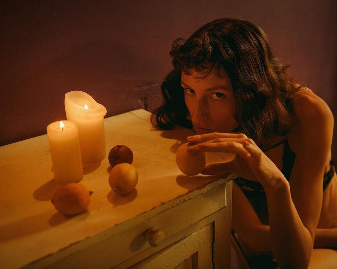 Woman with dark hair sitting at white table holding lemon with candles and fruit, creates a warm, vintage, and intimate ambiance.