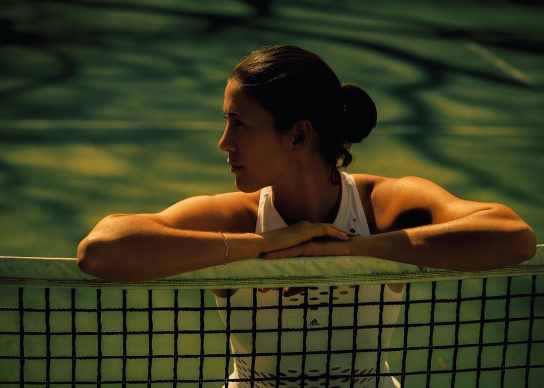 Woman poses on tennis court, resting arms on net. Close-up captures fitness style, with bright sunlight on her face and hair.