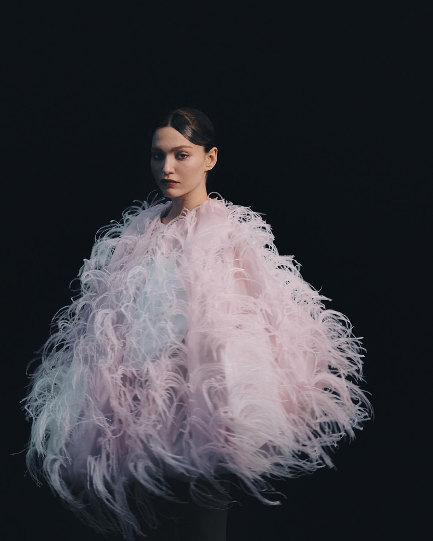 A striking portrait features a poised young woman in an extravagant, feathered pastel coat against a stark black background, studio lit.